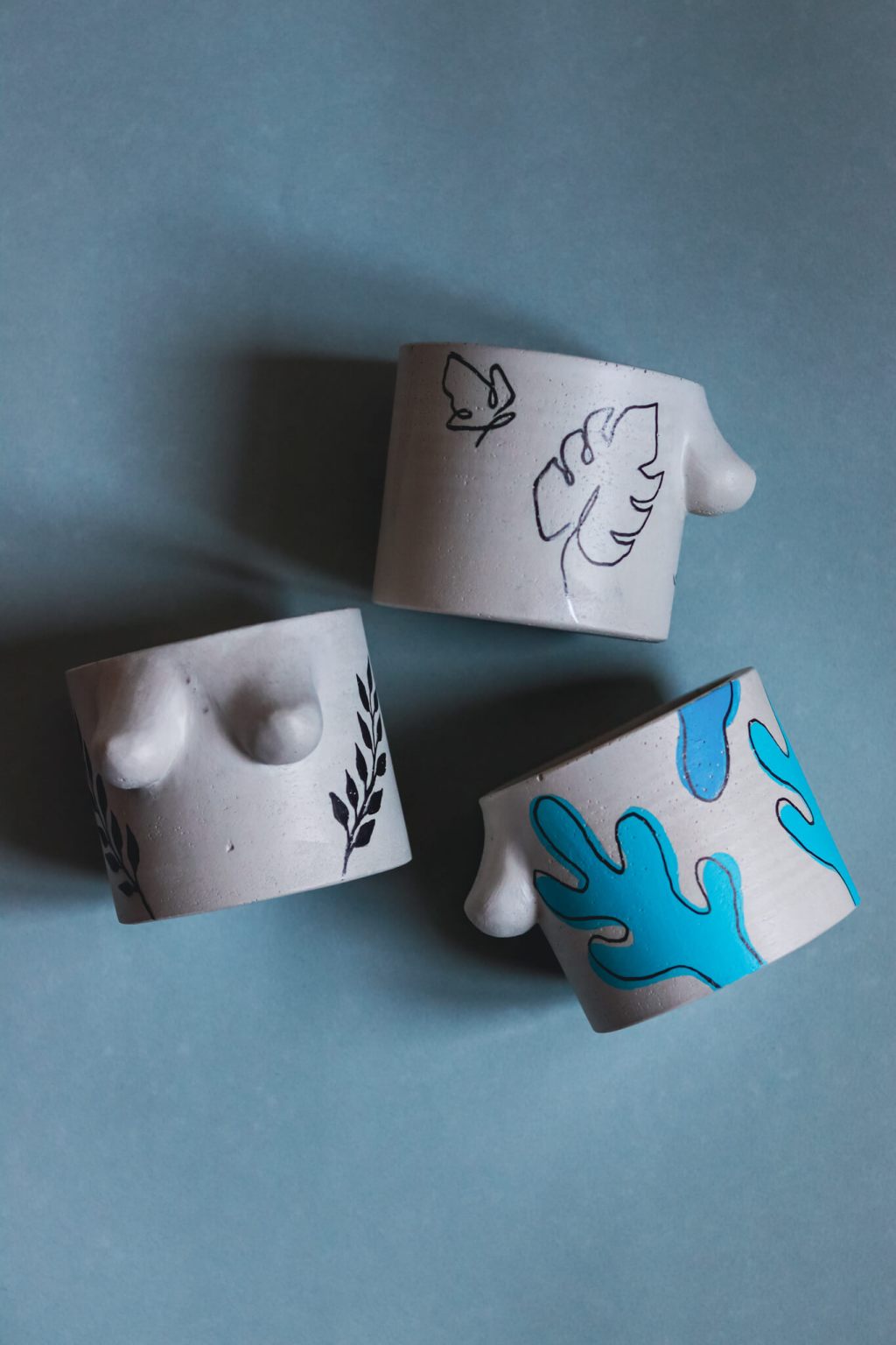 Drei handbemalte Boobie Pots von Viktoria Spokojna in Grau. Linker Pot mit schwarzen Pflanzensträngen, einem One-Line-Schmetterling und einem One-Line-Monsterablatt. Rechter Pot mit kaktusartigen Formen in schwarz und zwei Blautönen, die über den Rand gehen.