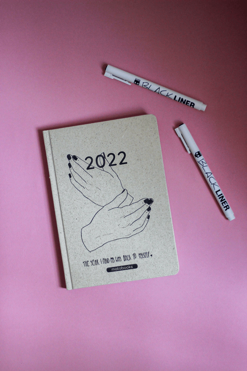 Photo of a matabooks blank calendar 2022 lying on a pink background, with two Skullpaper BLACK LINERs beside it. Drawing by Viktoria Spokojna: two hands showing the back of the hand, black nails, in a pose suggesting self-embrace. Text reads 